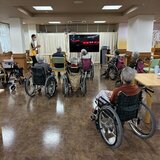 有料老人ホーム「スーパーコート高石羽衣」の食堂で、大画面テレビの前に車椅子の入居者様が集まり、スタッフと一緒に体操やカラオケのレクリエーションを楽しんでいる風景