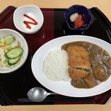 和風お盆に載った、スーパーコート高石羽衣の昼食「プライムメニュー」。白い楕円皿に盛られたカツカレー（白ご飯と豚カツ3切れ）をメインに、野菜サラダ（レタス、きゅうり、コーン）の小鉢、らっきょうと福神漬けの小皿、イチゴソースがかけられたヨーグルトの小鉢、スプーン、箸が配置されています。木製テーブルの上に置かれています。