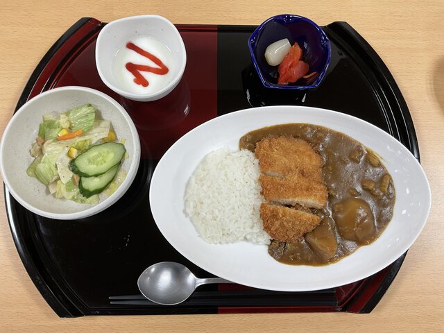 和風お盆に載った、スーパーコート高石羽衣の昼食「プライムメニュー」。白い楕円皿に盛られたカツカレー（白ご飯と豚カツ3切れ）をメインに、野菜サラダ（レタス、きゅうり、コーン）の小鉢、らっきょうと福神漬けの小皿、イチゴソースがかけられたヨーグルトの小鉢、スプーン、箸が配置されています。木製テーブルの上に置かれています。