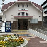 赤い屋根とステンドグラスが特徴的な南海高師浜線「高師浜駅」の駅舎。手前には綺麗に整備された花壇と、右側にはバリアフリーのスロープが映っている。