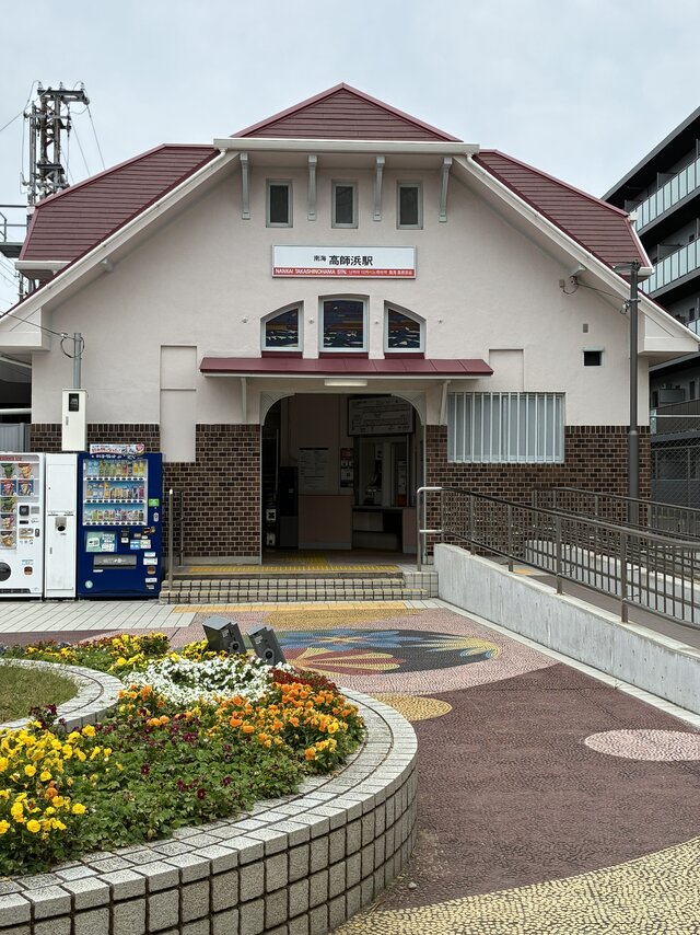 赤い屋根とステンドグラスが特徴的な南海高師浜線「高師浜駅」の駅舎。手前には綺麗に整備された花壇と、右側にはバリアフリーのスロープが映っている。