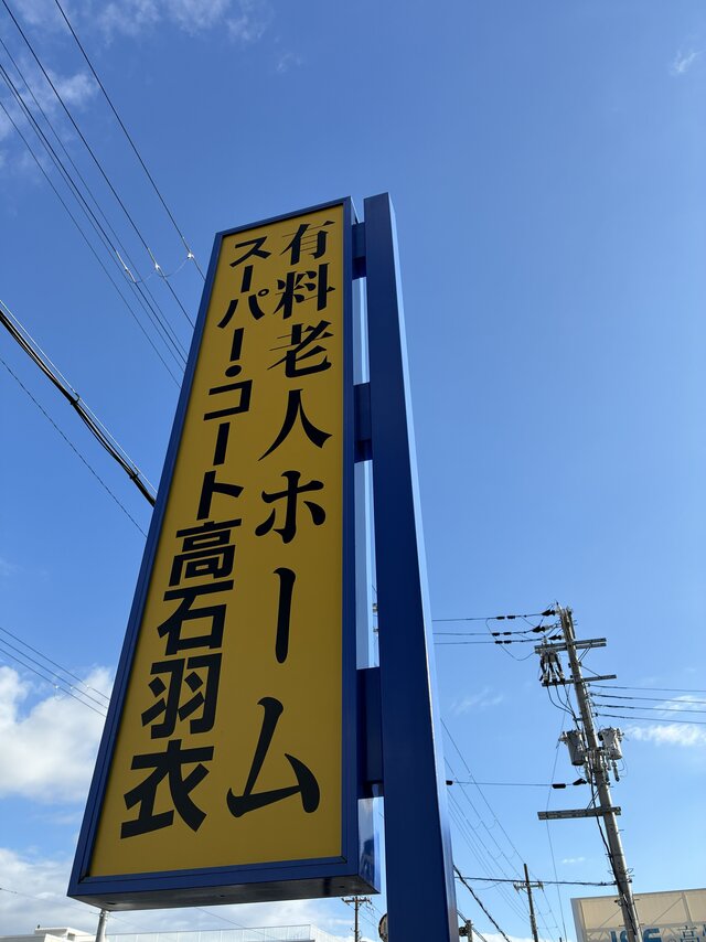 alt="青空を背景にした、有料老人ホーム スーパー・コート高石羽衣の黄色く大きな道路面看板"
