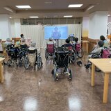 スーパーコート高石羽衣の広々とした食堂で、大型モニターに映るインストラクターに合わせて、車椅子や椅子に座ったご入居者様たちが一緒に手を動かして体操をしている様子