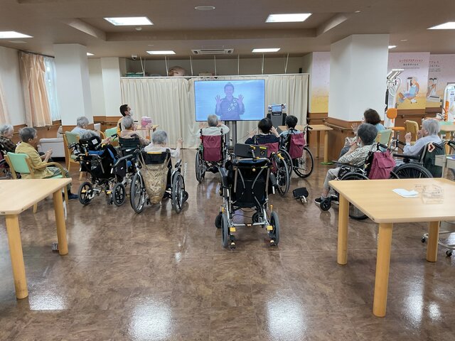 スーパーコート高石羽衣の広々とした食堂で、大型モニターに映るインストラクターに合わせて、車椅子や椅子に座ったご入居者様たちが一緒に手を動かして体操をしている様子