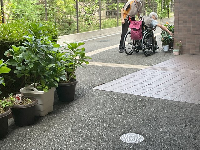 スーパーコート高石羽衣の敷地内で、車椅子に乗ったご入居者がスタッフに見守られながら、ハイビスカスの鉢植えに一生懸命手を伸ばして水やりをしている様子
