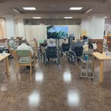 スーパーコート高石羽衣の広々としたフロアで、体操の後に大きなモニターを見ながら車椅子や椅子に座ってカラオケレクリエーションを楽しむ入居者様たちの後ろ姿。