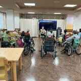 スーパーコート高石羽衣の広々とした食堂で、大きなモニターを囲むように車椅子や椅子に座り、スタッフと一緒に体操やカラオケのレクリエーションを楽しむ高齢者の入居者様たちの様子