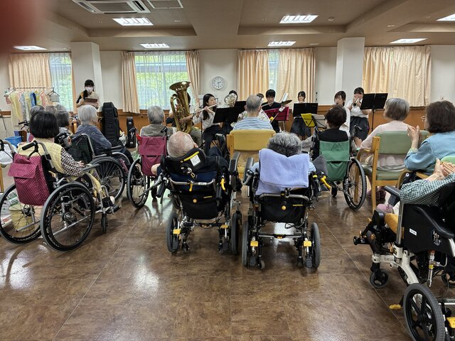 alt="スーパーコート高石羽衣の食堂にて、車椅子や椅子に座って信太高校吹奏楽部による楽器演奏を熱心に鑑賞する高齢の入居者様たちの様子"