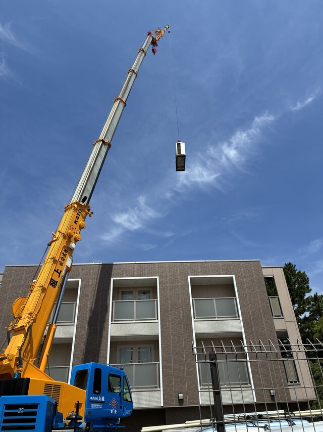 スーパーコート高石羽衣の建物前に配置された黄色い大型クレーン車と吊り上げられるエアコン室外機