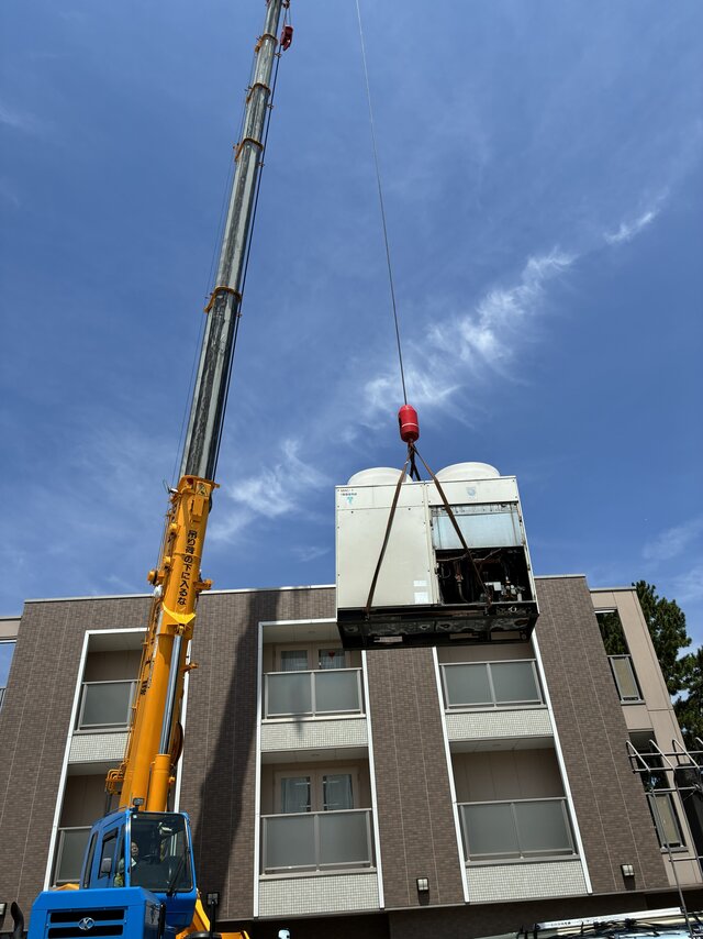 青空をバックに大型クレーン車で高く吊り上げられている大型エアコン室外機の様子