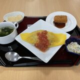 スーパーコート高石羽衣のイベントランチ「マルタマデー」のメニュー。フレッシュトマトソースがかけられたオムライスをメインに、海老カツ(ソース付き)、コンソメスープ、コールスローサラダ、クラッカーのような焼き菓子が黒いトレイに並べられた様子。