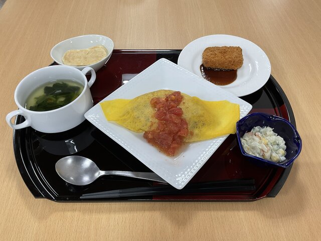 スーパーコート高石羽衣のイベントランチ「マルタマデー」のメニュー。フレッシュトマトソースがかけられたオムライスをメインに、海老カツ（ソース付き）、コンソメスープ、コールスローサラダ、クラッカーのような焼き菓子が黒いトレイに並べられた様子。