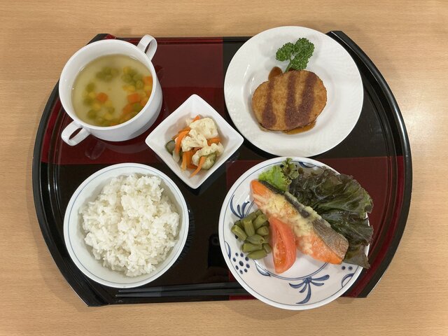 alt="スーパーコート高石羽衣の夕食「プライムメニュー」。銀鮭の和風チーズ焼き、コロッケ、カリフラワーのマリネ、コンソメスープ、ご飯が黒と赤のお盆にきれいに並べられた写真"