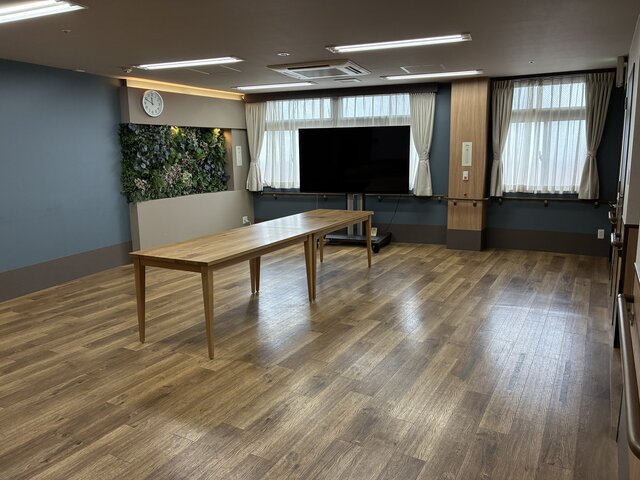 alt="スーパーコート高石羽衣の広々とした談話室。中央に置かれた木製の長テーブル、奥の大画面テレビ、壁面を彩るウォールグリーン、明るく清潔感のあるフローリングの室内"
