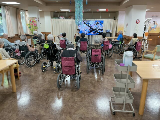 alt="スーパーコート高石羽衣の食堂で、正面のモニターとスタッフに向かって車椅子の入居者様たちが午後のタオル体操を行っている様子"