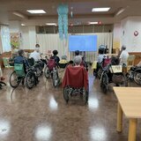 スーパーコート高石羽衣1のデイルームで、大型モニターを見ながら車椅子や椅子に座って集団体操を行う入居者様の様子