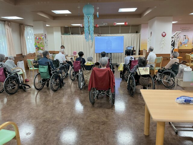 スーパーコート高石羽衣1のデイルームで、大型モニターを見ながら車椅子や椅子に座って集団体操を行う入居者様の様子