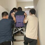 スーパーコート高石羽衣で行われた避難訓練の様子。職員3名が車いすに乗った入居者（を想定した職員）を協力して持ち上げ、狭い非常階段を上へと運んでいる。真剣な表情で連携を確認している。