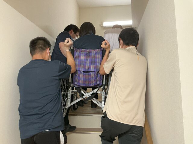 スーパーコート高石羽衣で行われた避難訓練の様子。職員3名が車いすに乗った入居者(を想定した職員)を協力して持ち上げ、狭い非常階段を上へと運んでいる。真剣な表情で連携を確認している。