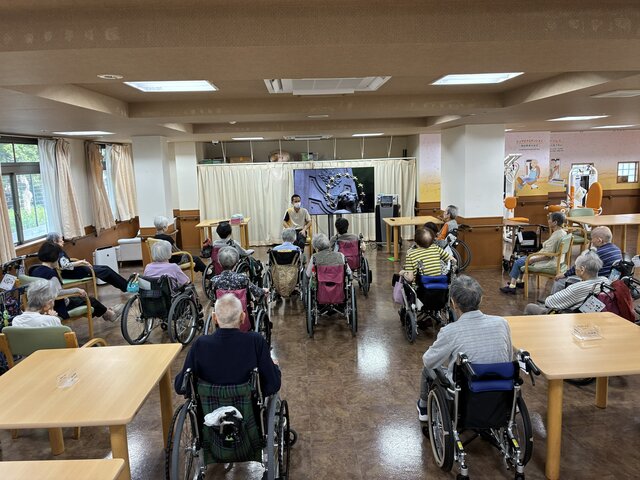 スーパーコート高石羽衣で昨日行われたストレッチ体操の様子。デイルームにて、正面のモニターとスタッフに合わせてご入居者が無理のない姿勢で体を動かしている風景。