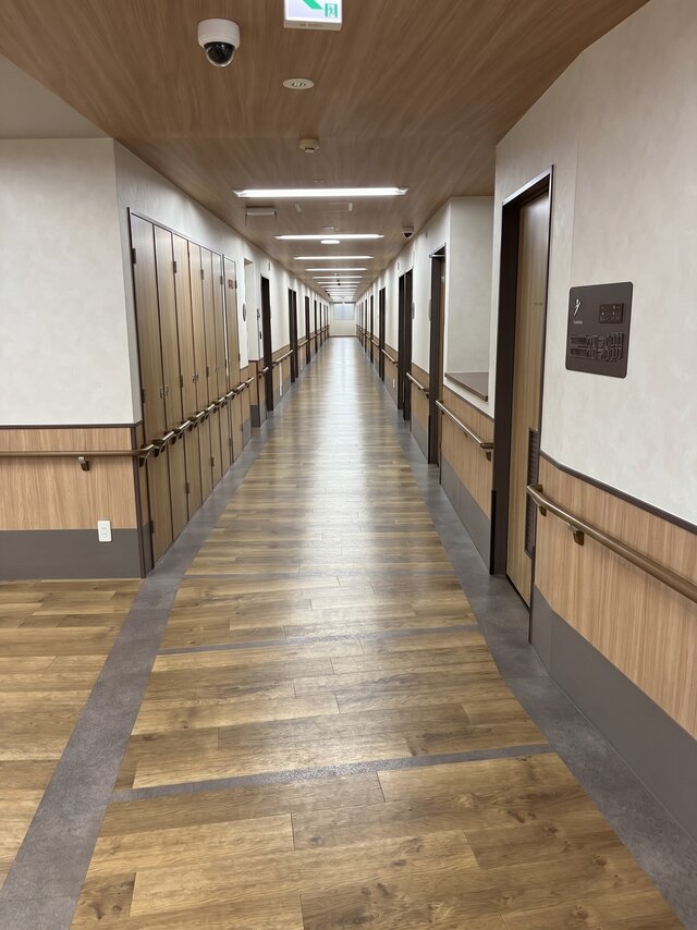 alt="大阪府高石市の高齢者住宅「スーパーコート高石羽衣」3階。手すりが完備された、木目調床の清潔で長い廊下の内観。"