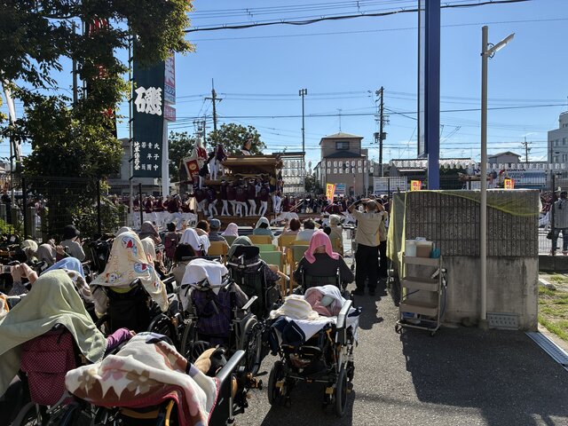 alt="スーパーコート高石羽衣の敷地近くで高石神社のだんじり祭り（宮入）を車椅子で見学するご入居者様の様子"
