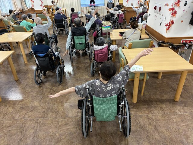alt="スーパー・コート高石羽衣のフロアで、モニターを見ながら車椅子に座ったご入居者様が腕を大きく広げて体操を行っている様子"